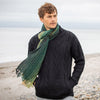 A man standing by the sea wearing a black knit sweater and a green alpaca wool scarf with a geometric pattern.