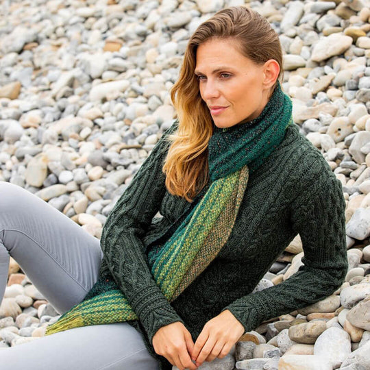 A woman sitting on a pebble beach wearing a green and brown alpaca wool scarf.