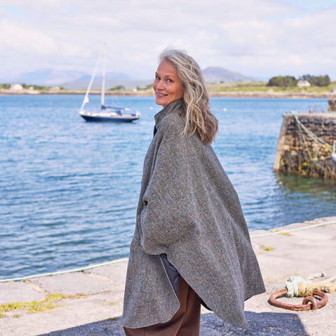 A woman with long, wavy gray hair stands by a waterfront wearing a gray cape. She smiles while looking back over her shoulder. In the background, a sailboat is visible on the water, along with a rocky shoreline and distant hills under a partly cloudy sky.