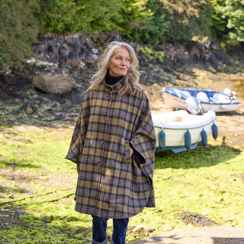 A woman with long, wavy hair stands on a rocky shoreline wearing a plaid, oversized cape. She gazes thoughtfully towards a small boat moored nearby, surrounded by greenery and water.