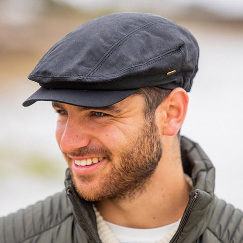 Black waxed Irish flat cap with subtle stitching and a small brass logo tag, worn outdoors with a quilted jacket.