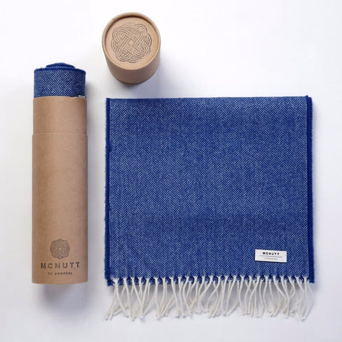 Blue herringbone lambswool scarf with cream fringe, shown folded beside its cylindrical McNutt packaging.