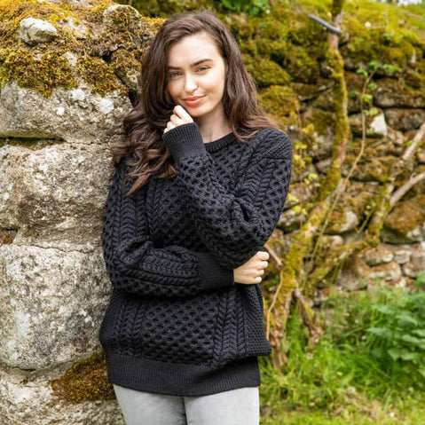 A young woman stands against a stone wall covered in moss. She is wearing a black Aran sweater with a textured pattern and light gray pants. The background features greenery, suggesting an outdoor setting.