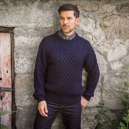 A man wearing a traditional crew neck Aran sweater in black, standing against a rustic background.
