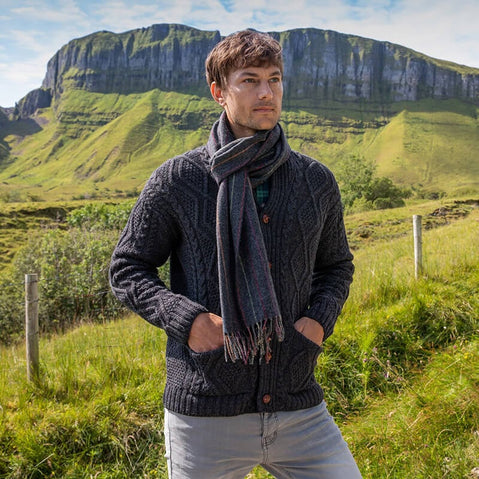 A man stands in a green landscape, wearing a dark knitted aran cardigan and a gray scarf. He has his hands in the pockets of his cardigan, with hills and a cliff visible in the background under a clear sky.