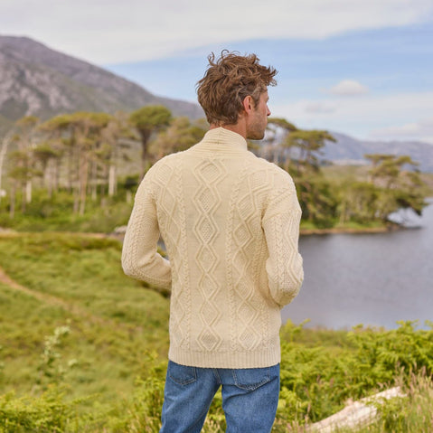A man stands with his back to the camera, wearing a cream-colored cable-knit aran sweater and blue jeans. He overlooks a serene landscape featuring a lake surrounded by lush greenery and mountains in the background under a clear sky.