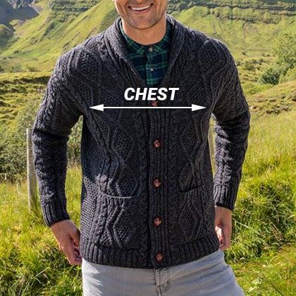 A man stands outdoors, smiling while wearing a dark gray knit aran cardigan with buttons. He is positioned in front of a lush green landscape, and the word