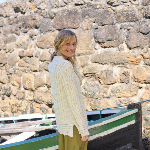 Person wearing a cream-colored Women's Signature Aran Tunic Sweater from The Irish Store, featuring traditional Aran stitching and a green skirt, standing in front of a stone wall with a green and white striped boat in the background