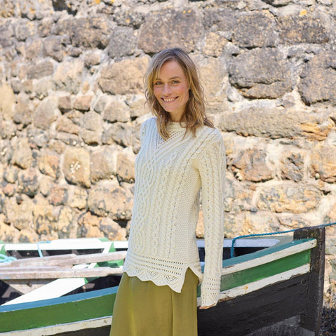 Person wearing a cream-colored Women's Signature Aran Tunic Sweater from The Irish Store, featuring traditional Aran stitching and a green skirt, standing in front of a stone wall with a green and white striped boat in the background