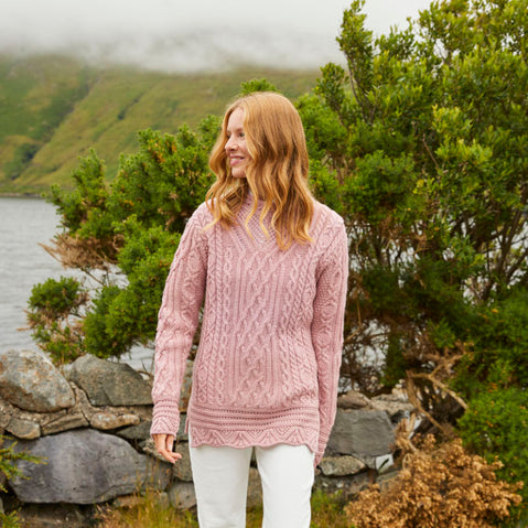 A woman standing in front of a stone wall wearing a beige Aran tunic sweater with a turtleneck design and scalloped edge at the hem.