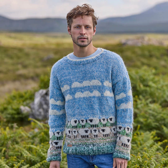A man wearing a hand-knitted blue sweater with a white sheep pattern standing in a field.