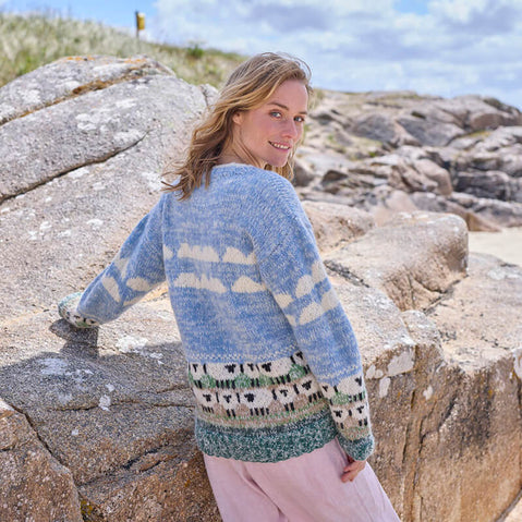 A woman standing by rock wearing a hand-knitted grey and white sheep patterned sweater.