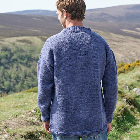 Man wearing a blue knitted sweater standing on a grassy hill with a scenic view.