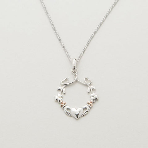 A silver pendant featuring a heart, shamrock, and claddagh design with rose gold accents on a chain.