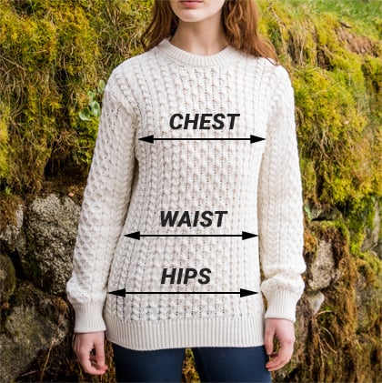 A person stands in front of a mossy stone wall, wearing a white knitted aran sweater. On the sweater, arrows indicate measurements labeled