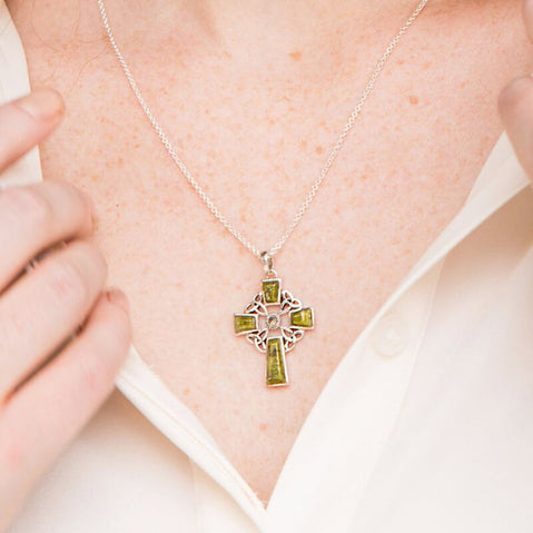A close-up of a person's neckline featuring a silver necklace with a green marble and silver Celtic cross pendant. The individual is adjusting their collar, revealing the pendant against a light-colored shirt.