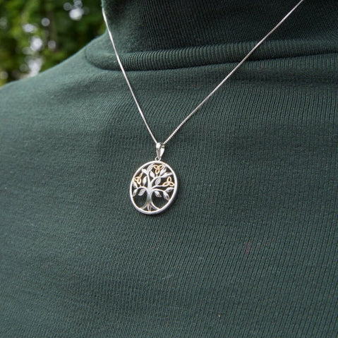 Silver Tree of Life pendant with gold‑tone trinity knot accents, worn on a fine chain over a dark green top outdoors.