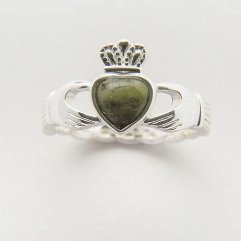 A silver Claddagh ring with a connemara marble stone at the center, placed on a rock with flowers in the background.