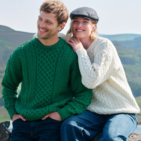 Man and woman wearing Aran sweater and cap sitting outdoors with mountains in the background