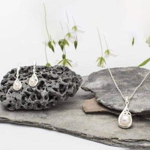 Silver Trinity Knot pearl jewelry set featuring drop earrings and a matching pendant, displayed on textured stone with soft white flowers in the background.