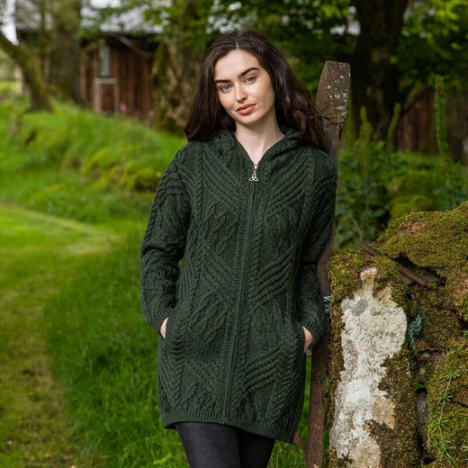A woman standing by a rustic fence wearing a green Aran zipper coat with a hood, featuring a trinity knot design.