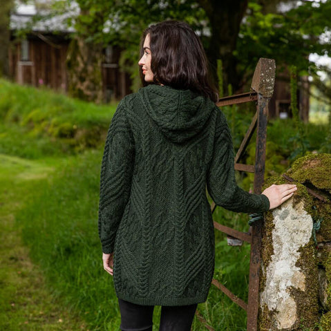A woman with shoulder-length dark hair stands with her back to the camera, wearing a dark green Aran cardigan. She faces a grassy area with trees in the background and is holding onto a rustic gatepost. The setting appears serene and natural.