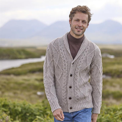 A man wearing a gray Aran shawl collar button cardigan with a geometric pattern, standing on a rock with a scenic landscape in the background.