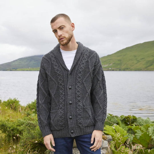 Man wearing a gray knitted cardigan standing by a lake with mountains in the background