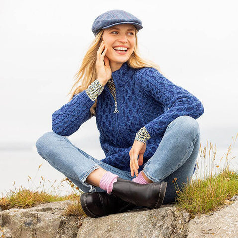 A woman is sitting on a rocky surface, smiling and wearing The Kenmare Aran Cardigan in blue, and a gray cap. She has her hand resting on her cheek and is casually dressed with rolled-up jeans and black boots. The background features a cloudy sky and greenery, suggesting an outdoor setting.