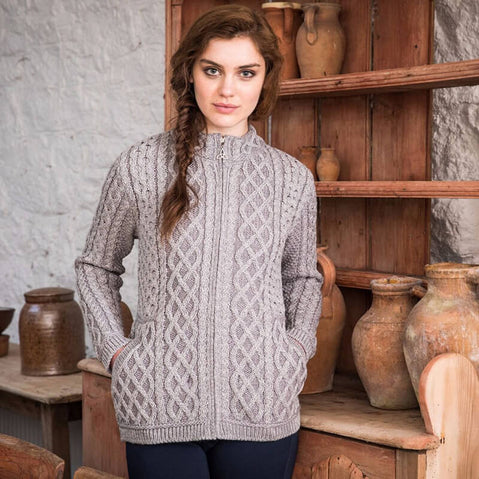 A young woman stands confidently in front of a rustic wooden shelf adorned with clay pots. She is wearing a textured grey aran cardigan with a high collar and pockets, styled simply with dark pants. The background features stone walls, emphasizing a warm, cozy atmosphere.
