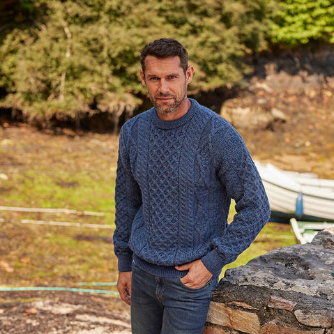 A man stands outdoors, wearing a textured blue aran sweater and dark jeans. He poses casually near a stone wall, with a natural landscape of trees and boats in the background. The setting appears tranquil and rustic.