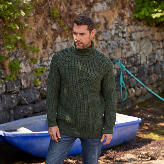 A man stands in front of a stone wall wearing an Irish fisherman sweater made in Ireland . A small blue boat is visible nearby, with greenery in the background suggesting an outdoor setting.