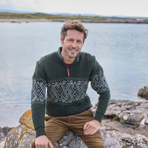 A man sitting on a rock wearing a green aran sweater with Celtic knot patterns, smiling at the camera with a body of water and landscape in the background.