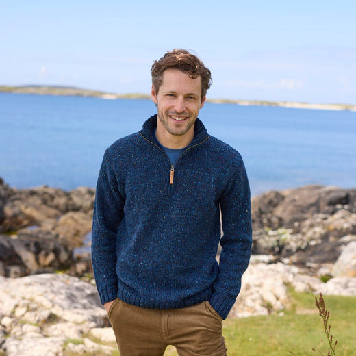 Man wearing a charcoal men's half zip fleck sweater and brown pants, posing by a rocky coast with ocean waves and distant hills in the background