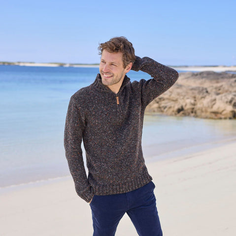 Man standing on a beach wearing a charcoal men's half zip fleck sweater, with a relaxed pose against a scenic ocean and rocky backdrop