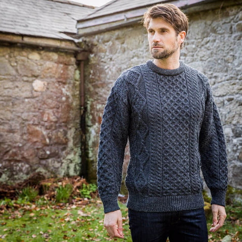 A man stands outdoors against a stone wall, wearing a dark cable-knit aran sweater and jeans. The setting is surrounded by grass and leaves, suggesting an autumn or early winter scene.