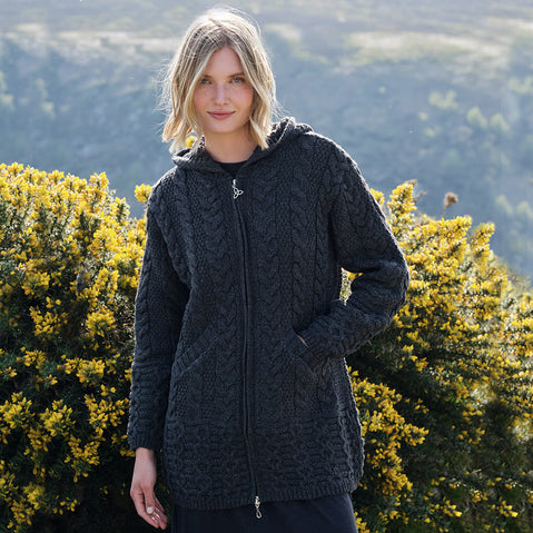 Woman wearing a dark gray knitted hoodie standing outdoors with greenery in the background