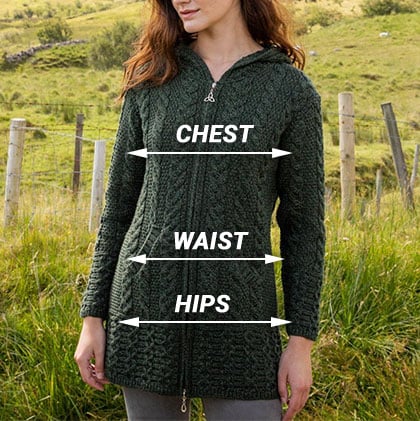 A woman stands in a grassy field, wearing a green knitted aran cardigan with a hood. Lines point to her chest, waist, and hips, indicating the measurements for sizing. The background features rolling hills and wooden posts.