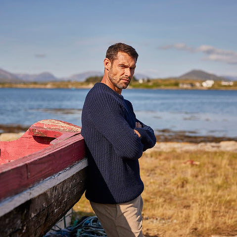 A man stands leaning on a boat by the coast wearing a traditional Irish fisherman sweater.