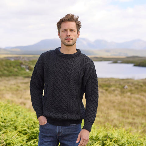 A man stands outdoors, wearing a dark cable-knit aran sweater and jeans. He is positioned in front of a scenic landscape with a small lake and rolling hills in the background under a partly cloudy sky.