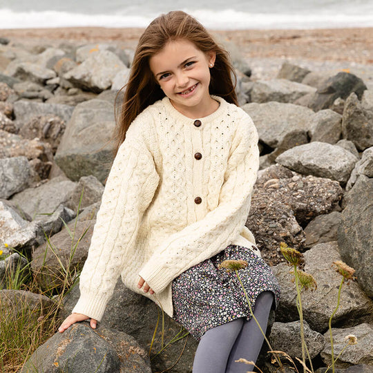 Girl sitting on a rocky beach waering a cream merino wool Kids Aran cardigan with traditional cable knit patterns and brown buttons
