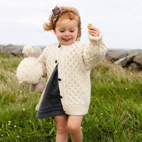 Child playing in a field wearing cream kids Aran sweater with traditional Irish knitwear patterns. She holds a yellow flower and a teddy bear
