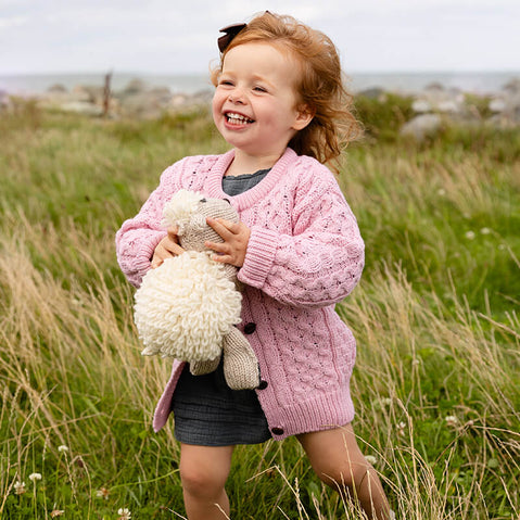 Child running in a grassy field holding a teddy bear wearing a pink kids aran merino wool cardigan with traditional Irish cable and honeycomb stitches