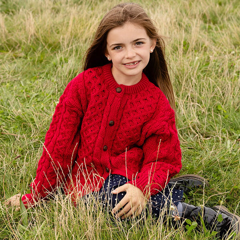 A young girl sits in a grassy field, wearing a bright red knitted aran cardigan with buttons. She has long brown hair and smiles at the camera, surrounded by tall grass.
