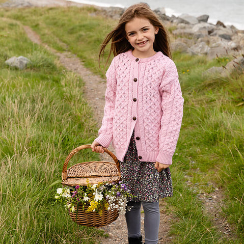 A young girl stands in a field by the coast ,wearing a pink knitted kids Aran cardigan with buttons. She has long brown hair and smiles at the camera holding a basket of wildflowers.