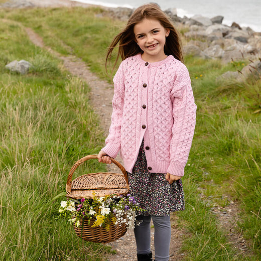 A young girl stands in a field by the coast ,wearing a pink knitted kids Aran cardigan with buttons. She has long brown hair and smiles at the camera holding a basket of wildflowers.