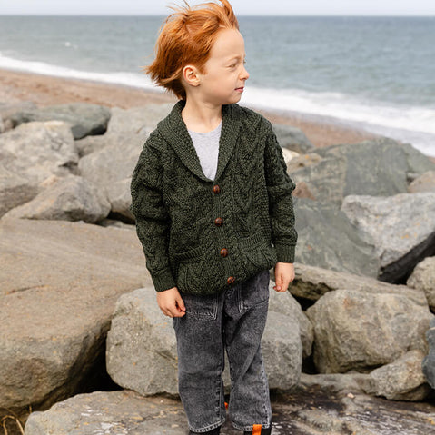 A young child with red hair stands on a rocky beach, wearing a dark green aran sweater and gray pants. The ocean can be seen in the background, with gentle waves lapping at the shore.