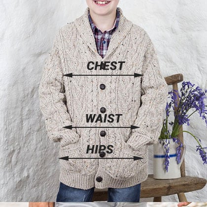 A smiling child stands against a textured white backdrop, wearing a beige, knitted cardigan. Measurement lines labeled