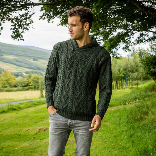 A man stands outdoors in a green cable-knit aran sweater and gray jeans, gazing slightly off to the side. He is amidst a grassy landscape with rolling hills in the background under a partly cloudy sky. Trees are visible nearby.