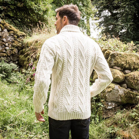 A man stands with his back to the camera, wearing a cream-colored cable-knit aran sweater. He is outdoors, surrounded by greenery and stone walls. The image captures a serene, natural setting.